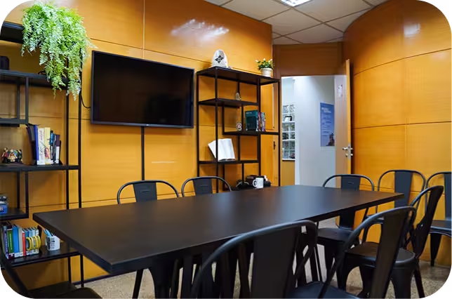 A imagem mostra uma sala moderna de reuniões com uma mesa preta central, cercada por cadeiras. Ao fundo, há estantes de metal com livros e uma planta decorativa, além de uma TV na parede e uma porta de acesso à outra área. O ambiente é acolhedor e bem iluminado.