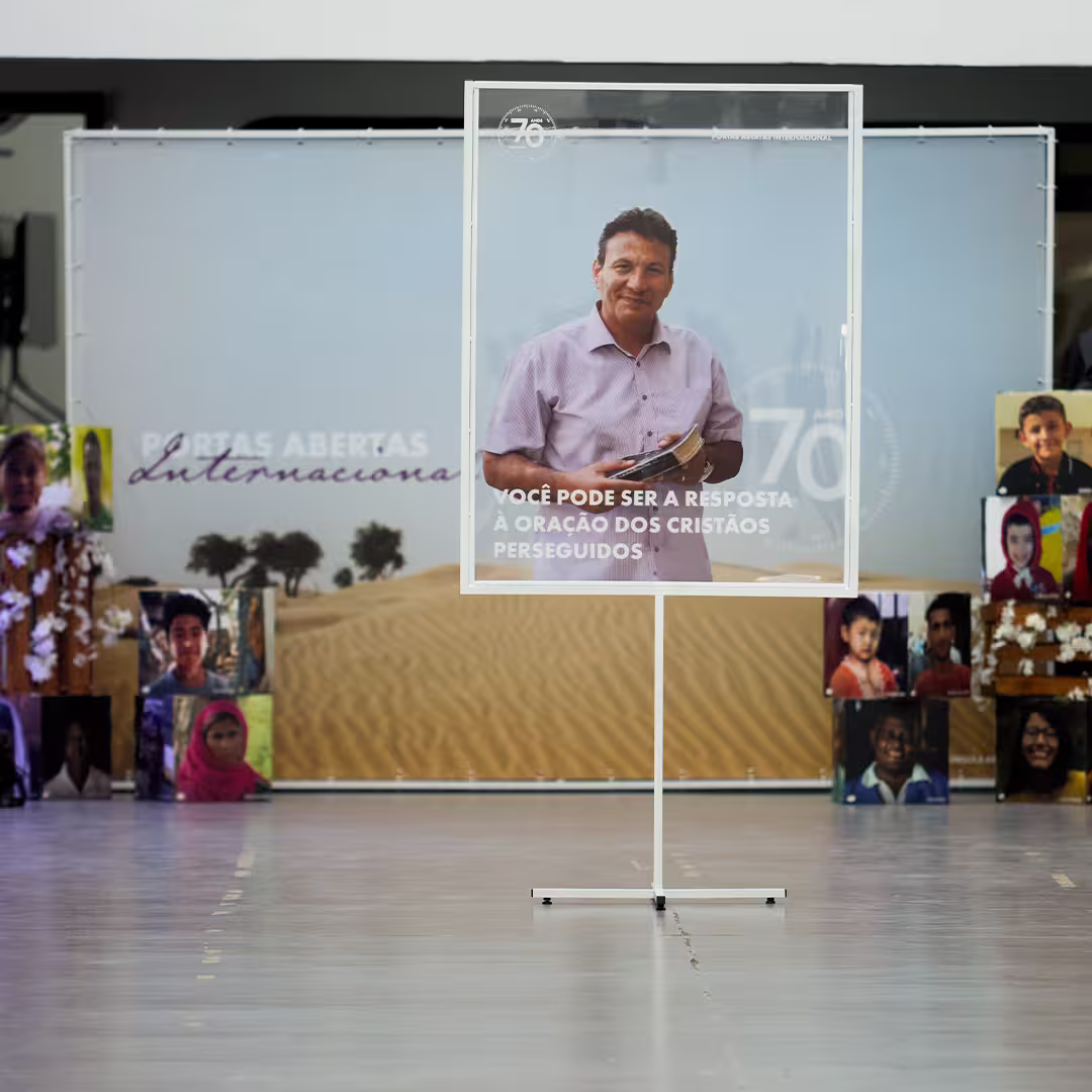 O Irmão André sorri ao lado de um amigo em um ambiente descontraído, ambos sentados em uma mesa com livros e papéis. O ambiente ao fundo sugere um momento de convivência e trabalho em grupo, com outras pessoas participando de atividades. Ambos parecem acolhedores e envolvidos em um contexto de parceria e fé.