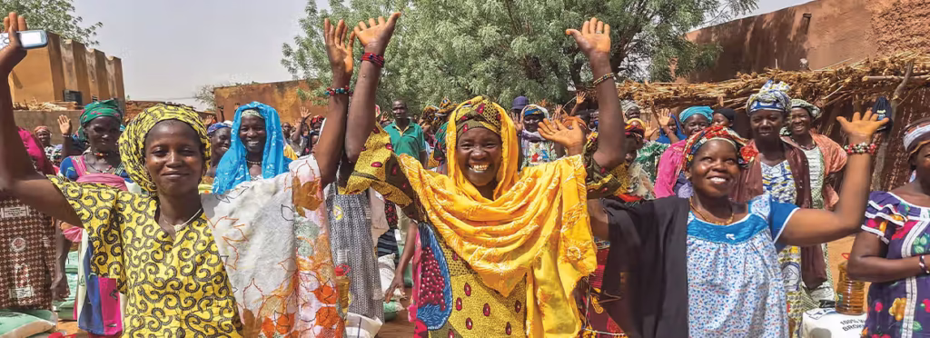 Grupo de cinco pessoas posando ao ar livre em uma área rural, com árvores e construções simples ao fundo. Um homem veste camiseta listrada amarela e preta, enquanto quatro mulheres usam hijabs coloridos e seguram Bíblias. Todos olham para a câmera com expressões calmas e amigáveis, sugerindo um momento de união ou atividade comunitária.