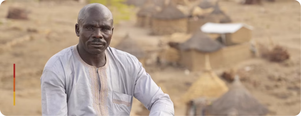 Homem com expressão séria posando ao ar livre, com árvores desfocadas ao fundo. Ele veste uma camisa branca com detalhe laranja. No canto inferior esquerdo da imagem, há uma barra colorida em tons de amarelo, laranja e vermelho.