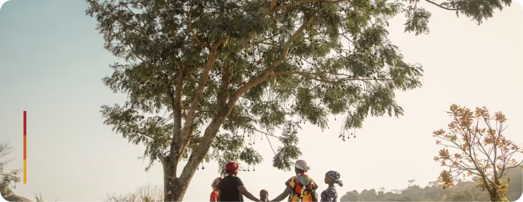 Homem com expressão de preocupação, vestindo camisa vermelha, posando em uma área verde com árvores desfocadas ao fundo. A luz do sol ilumina parcialmente seu rosto. No canto inferior esquerdo, há uma barra colorida em tons de amarelo, laranja e vermelho.