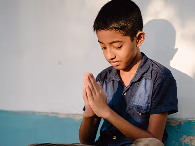 Mulher vestida com roupa azul estampada e xale da mesma cor está com as mãos erguidas em posição de oração. Ela usa relógio e anéis dourados. Ao fundo, outras pessoas também estão com os olhos fechados e em atitude de oração, demonstrando um momento coletivo de oração.