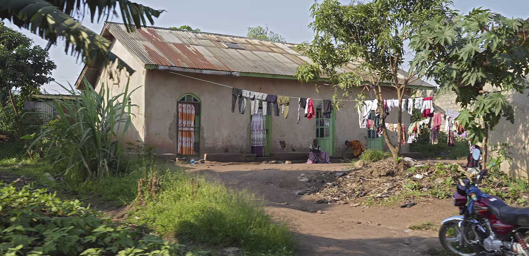 Casa na República Democrática do Congo. É possível ver pessoas sentadas à frente junto a um varal com roupas secando.
